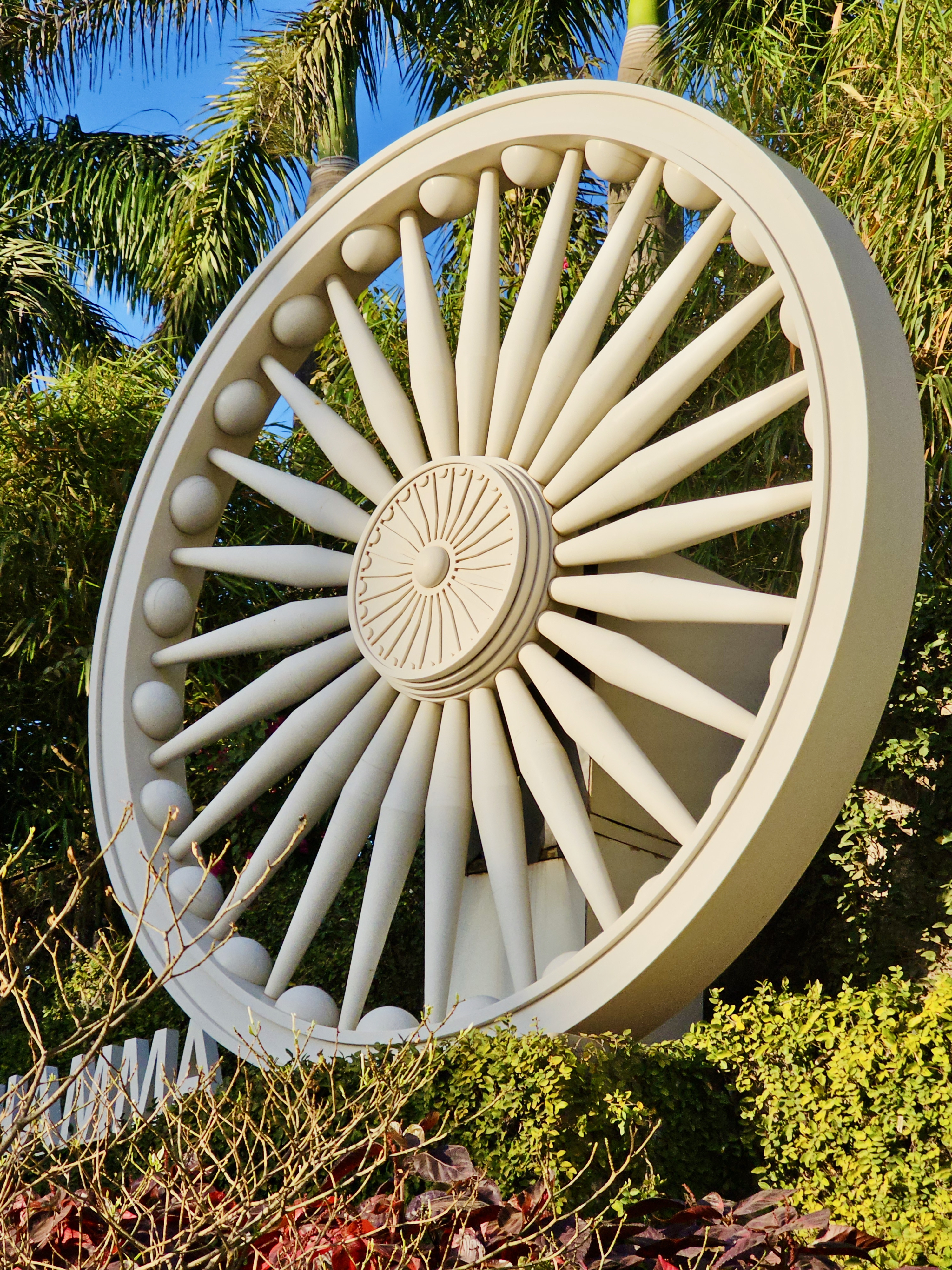 A large Ashoka Chakra wheel sculpture surrounded by greenery at Global Vipassana Pagoda, Gorai Village, Mumbai. The bright light and strong shapes make it visually striking.
