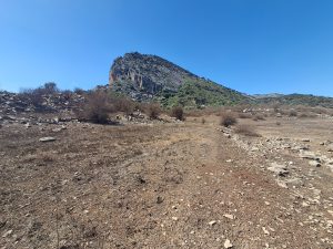 A dry, rocky landscape with sparse, low vegetation under a clear blue sky. In the background, a steep, rugged hill rises, partially covered with greenery. Montejaque (Málaga)