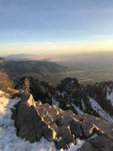 A scenic mountain view showing rocky terrain in the foreground with patches of snow. In the background, rolling hills and valleys extend towards the horizon, illuminated by the soft glow of a sunset