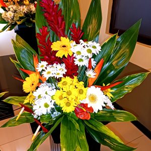 A vibrant floral arrangement featuring a variety of flowers, including red ginger, yellow sunflowers, white daisies, and colorful tropical blooms, complemented by lush green leaves