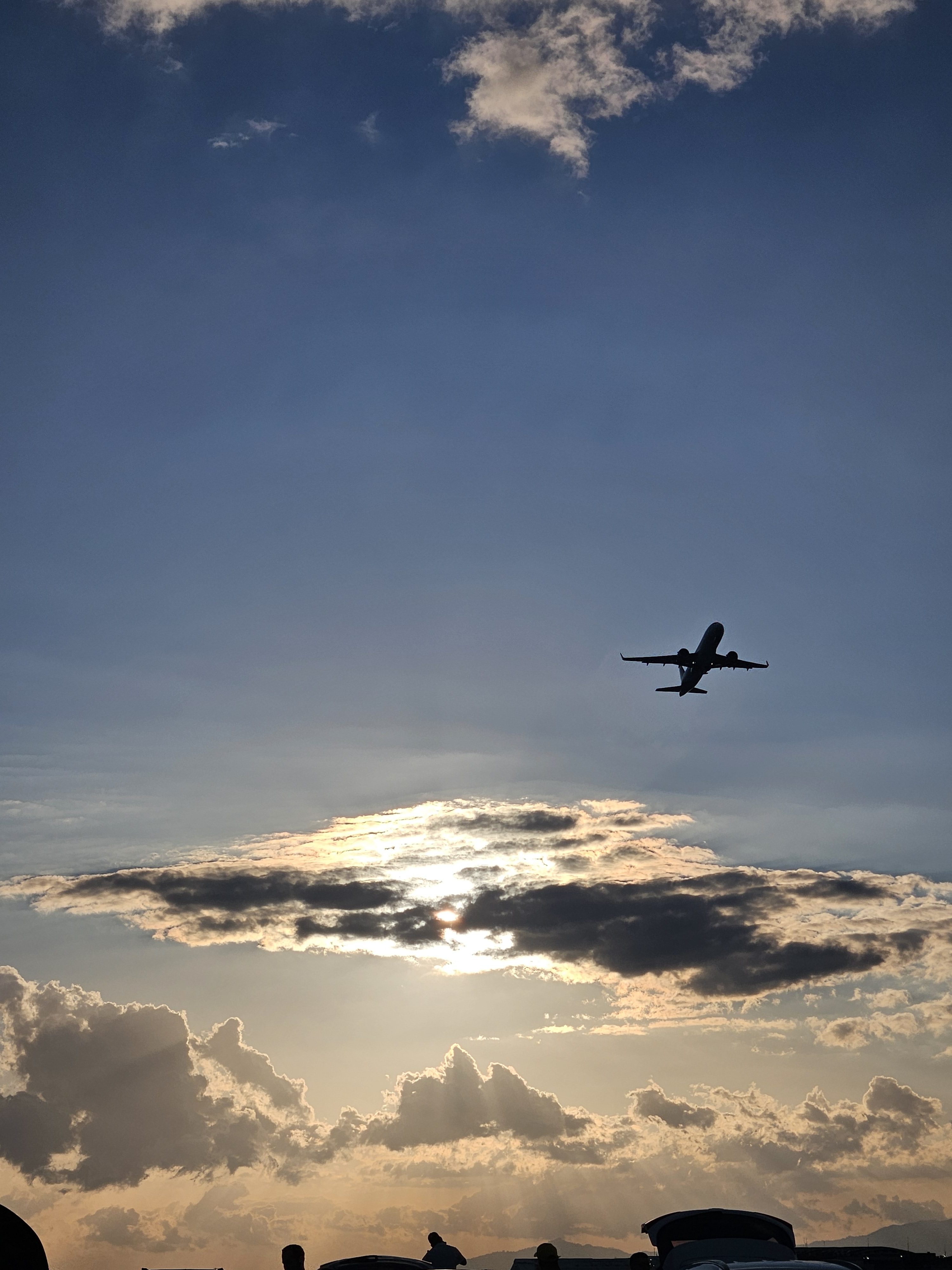 A silhouette of an airplane flying against a backdrop of a sunset. The sun is partially obscured by clouds, casting a golden glow in the sky.