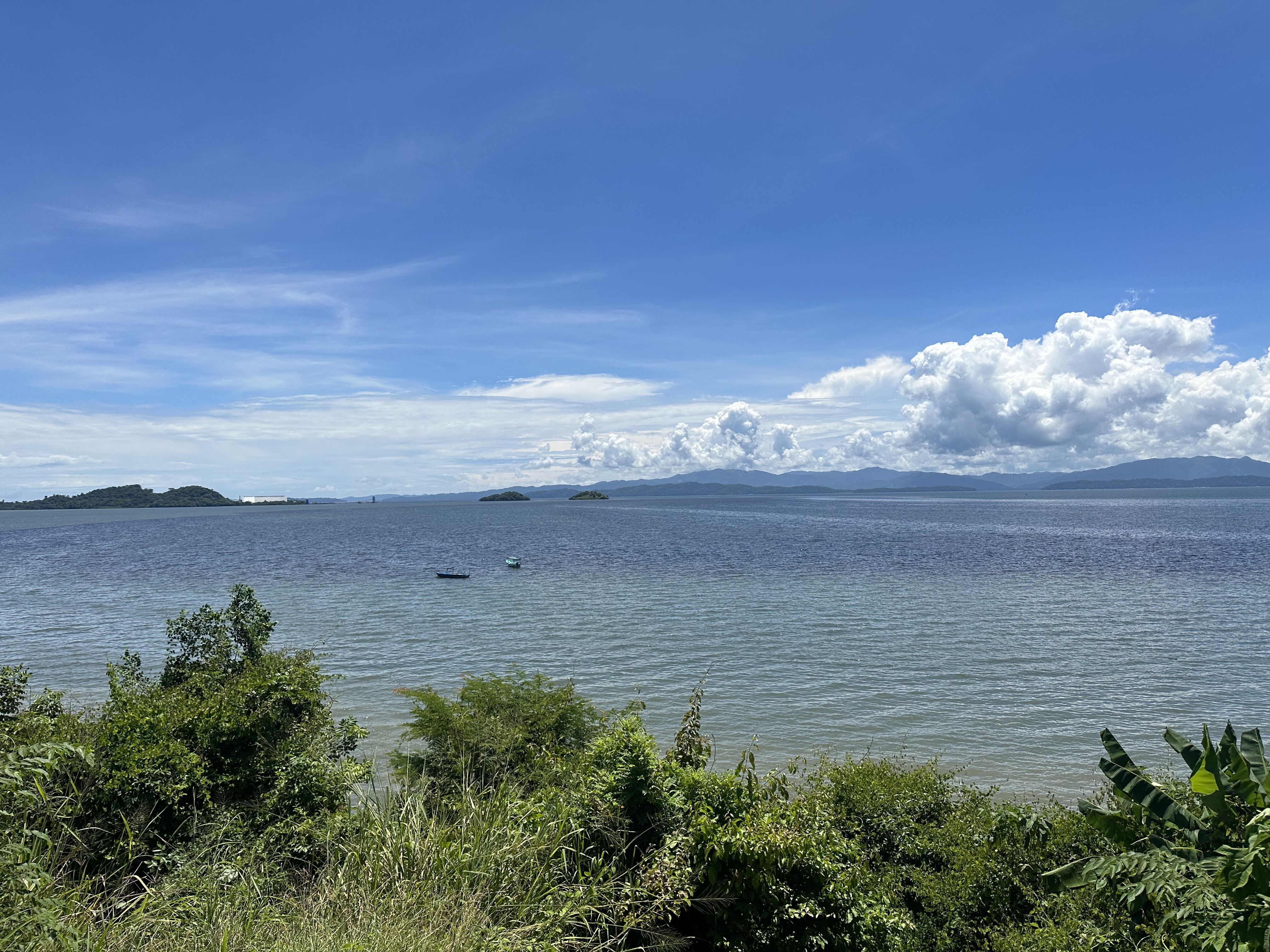 A scenic view of a calm sea under a bright blue sky with fluffy white clouds. In the distance, several small green islands can be seen on the horizon, along with a faint outline of land