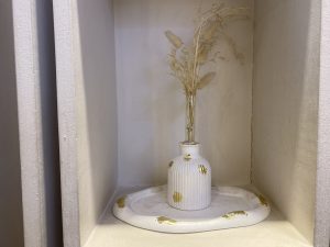 A white, ribbed vase with gold accents holds dried grass on a matching white and gold tray in a minimalist shelf. The scene feels calm and elegant.