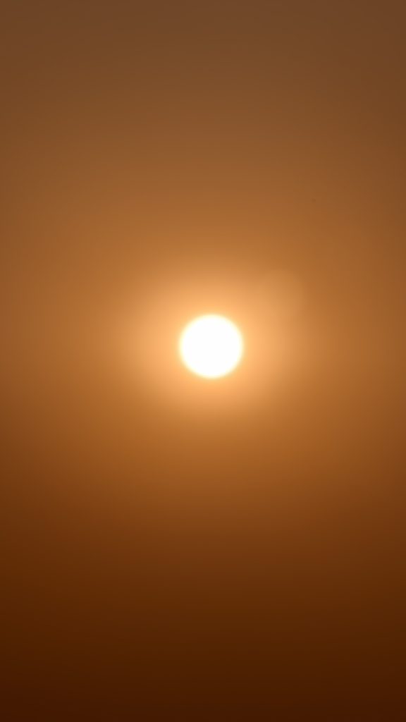 A minimalist vertical shot of a bright white sun centered in a solid orange and hazy sky.