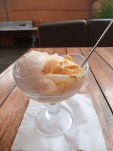 A glass bowl filled with three scoops of ice cream rests on a wooden table. The ice cream includes a creamy white flavor, a light yellow flavor, and a more vibrant yellow flavor. 