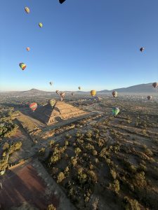 A view from above shows many colorful hot air balloons in a clear blue sky. Below, the Pyramid of the Sun stands out, surrounded by green land and some dry areas.
