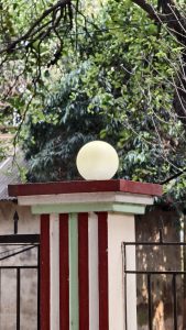 A round white light globe sits on top of a red and white striped concrete pillar next to a black metal fence with green trees behind it.