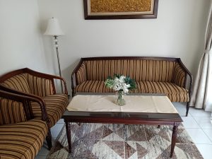 A cozy living room setting featuring two upholstered striped sofas with wooden frames. In the center, there's a glass-top coffee table with a beige cloth and a small vase filled with white flowers and green leaves