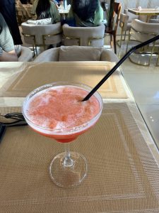 A frothy, pink cocktail in a glass with a black straw sits on a woven placemat. Plush chairs and people are visible in the background, creating a relaxed atmosphere.