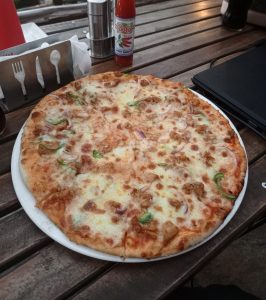 Thin-crust pizza with mozzarella, grilled chicken, bell peppers, and red onions on a white plate, served on a rustic wooden table with hot sauce and a napkin holder in daylight.