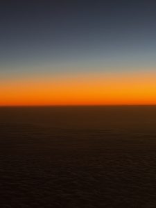A serene gradient sky at dusk, transitioning from a deep navy blue at the top to a vibrant orange along the horizon, with a smooth plane of clouds visible below, creating a tranquil atmosphere.