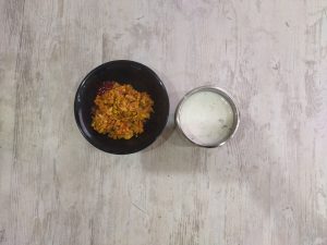 Two small bowls sit on a wooden table. One has a spiced egg and onion dish with a little ketchup, and the other contains creamy herb raita.