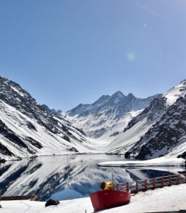 A scenic winter landscape featuring a tranquil, mirror-like lake surrounded by snow-covered mountains under a clear blue sky. In the foreground, a red boat is docked along the shoreline, while the dramatic peaks rise majestically in the background, hinting at the beauty of a crisp, sunny day. - Inca lake in Chile