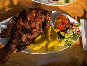 Deep-fried whole fish plated with potato wedges, mixed salad, and a side of spicy dipping sauce on a wooden table