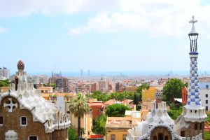 Panoramic view of Barcelona from Park Güell, featuring Gaudi's mosaic-tiled rooftops and spires in the foreground against the city skyline and Mediterranean Sea.