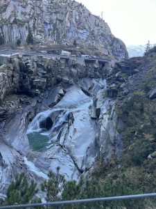 Rocky gorge with a waterfall flowing into a blue natural pool, with a road above through cliffs.