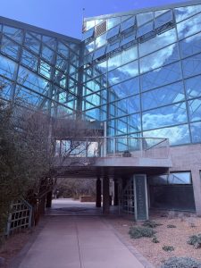 Botanical Gardens: a glass building/greenhouse for plants. The glass is reflecting the blue sky and wispy clouds. There is a walkway under one side of the building with a sign beside it that reads "Mediterranean Conservatory."