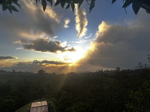 A picturesque landscape at sunset, featuring a dramatic sky filled with clouds in varying shades of blue, orange, and gray. Rays of sunlight break through the clouds, illuminating the horizon and casting a warm glow over a lush green hillside. In the foreground, a small wooden structure can be seen, partially obscured by greenery and trees, enhancing the serene natural setting.