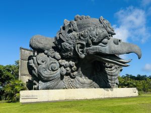 A large carved stone statue of a mythical bird with ornate details, set outdoors against a blue sky and greenery, with “PLAZA GARUDA” engraved at the base.
