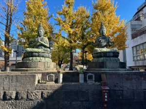 Two large bronze statues of seated Buddhas are positioned on a stone platform, surrounded by vibrant autumn trees with yellow leave