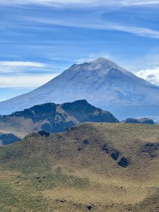 A massive volcanic cone dominates the background, its steep gray-brown slopes rising to a rounded summit shrouded in a thin white plume against a bright blue sky with wispy clouds. In the foreground, rolling hills of short golden-green grass lead to darker jagged ridges, with Popocatépetl — México’s iconic active volcano — towering behind them.
