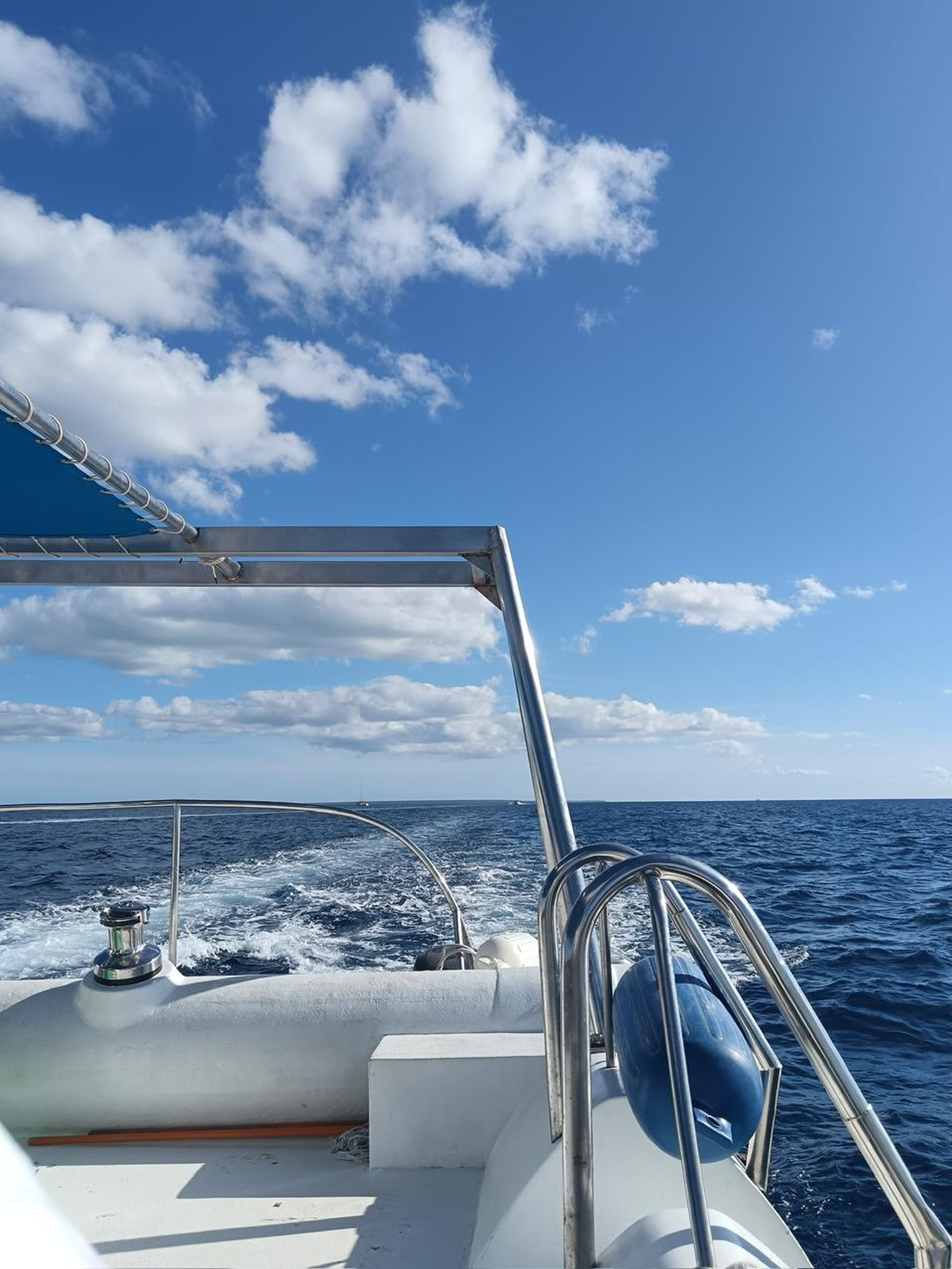 A view from the rear deck of a white boat sailing across a deep blue open ocean under a partly cloudy sky