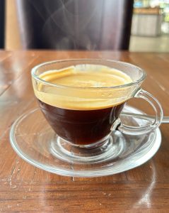 A glass cup filled with dark coffee sits on a clear glass saucer on a wooden table. 
