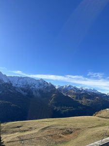 A panoramic view of a mountainous landscape under a clear blue sky. Snow-capped peaks rise majestically in the background, surrounded by green and yellow foliage on sloping hills. The foreground features grassy meadows with some patches of bare earth, creating a serene natural scene.