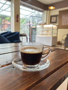 A clear glass cup of coffee sits on a wooden table in a cozy cafe, with a pastry display and warm lighting in the blurred background.