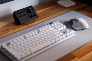 Close-up of a white mechanical keyboard and gaming mouse on a grey pad, with an audio control panel on a wooden desk in the background.