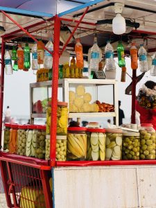 A vibrant food cart is displayed, featuring an array of glass jars filled with pickled fruits and vegetables, including bananas, cucumbers, and green balls. The cart is accentuated with colorful bottles of soft drinks and water hanging from the ceiling. 