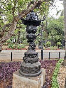 An artistic black metal lamp post with detailed carvings in a garden area in Chhatrapati Shivaji Maharaj Vastu Sangrahalaya, Mumbai. Surrounded by trees and plants, it adds a vintage charm. 
