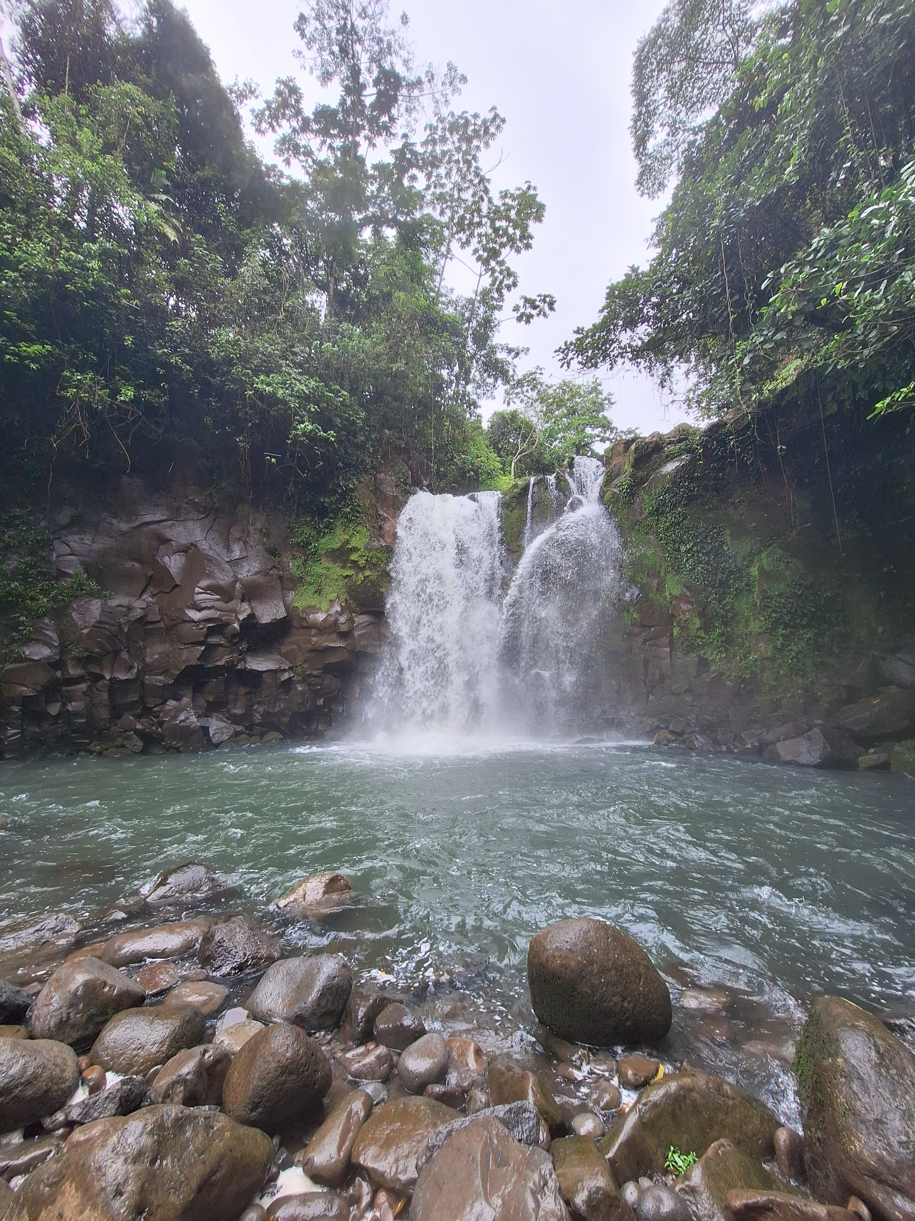 A waterfall cascades over rocky cliffs into a tranquil, greenish-blue pool surrounded by lush vegetation.