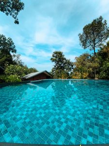 
An aerial view of a shimmering, turquoise tiled infinity pool surrounded by lush greenery and trees, under a bright blue sky with soft clouds