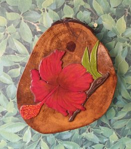 A wooden decoration shaped like a slice of tree trunk features a vibrant red hibiscus flower with a green leaf and a brown stem. The background is decorated with green leafy patterns, enhancing the natural theme of the piece. The hibiscus and the accompanying elements are artistically rendered, showcasing detail and color contrast against the natural wood texture.