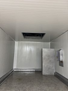An empty, clean room with smooth, white walls and a ceiling featuring a vent opening. The floor is gray and appears to be tiled. On one side of the room, there is a large, metallic cabinet, and on another wall, there is a small, metallic panel with an opening. The overall environment has a sterile, industrial look.