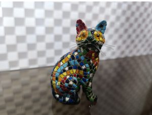 Escultura pequeña de un gato hecho de mosaicos de múltiples colores.(A small cat sculpture made of vibrant, multi-colored mosaics, standing on a neutral surface.)