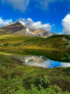 Asahidake, Hokkaido’s highest active volcano, rises steeply above lush alpine vegetation, its bare upper slopes streaked with white plumes of sulfurous steam. A still pond mirrors the peak and blue sky above. Low shrubs with small white flowers fill the foreground. Daisetsuzan National Park, Japan.
