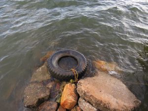 A weathered black tire sits partially submerged among large, jagged rocks at the edge of the shores of Lake victoria. Gentle ripples move across the greyish-green surface of the water around the rocks.