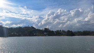 A panoramic view of a serene reservoir surrounded by green hills dotted with houses. A dramatic sky, filled with white and gray clouds, dominates the scene as the sun filters through gently, creating glints on the water. In the distance, a bridge connects the shores, completing a peaceful and picturesque landscape in the daylight.