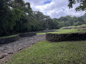 A view of an ancient stone structure surrounded by lush greenery and trees. The ground is covered in grass and stone pathways, leading to a prominent circular stone formation. In the background, there are additional stone remnants and a forested hillside under a partly cloudy sky. monumento nacional guayabo, parcelas 