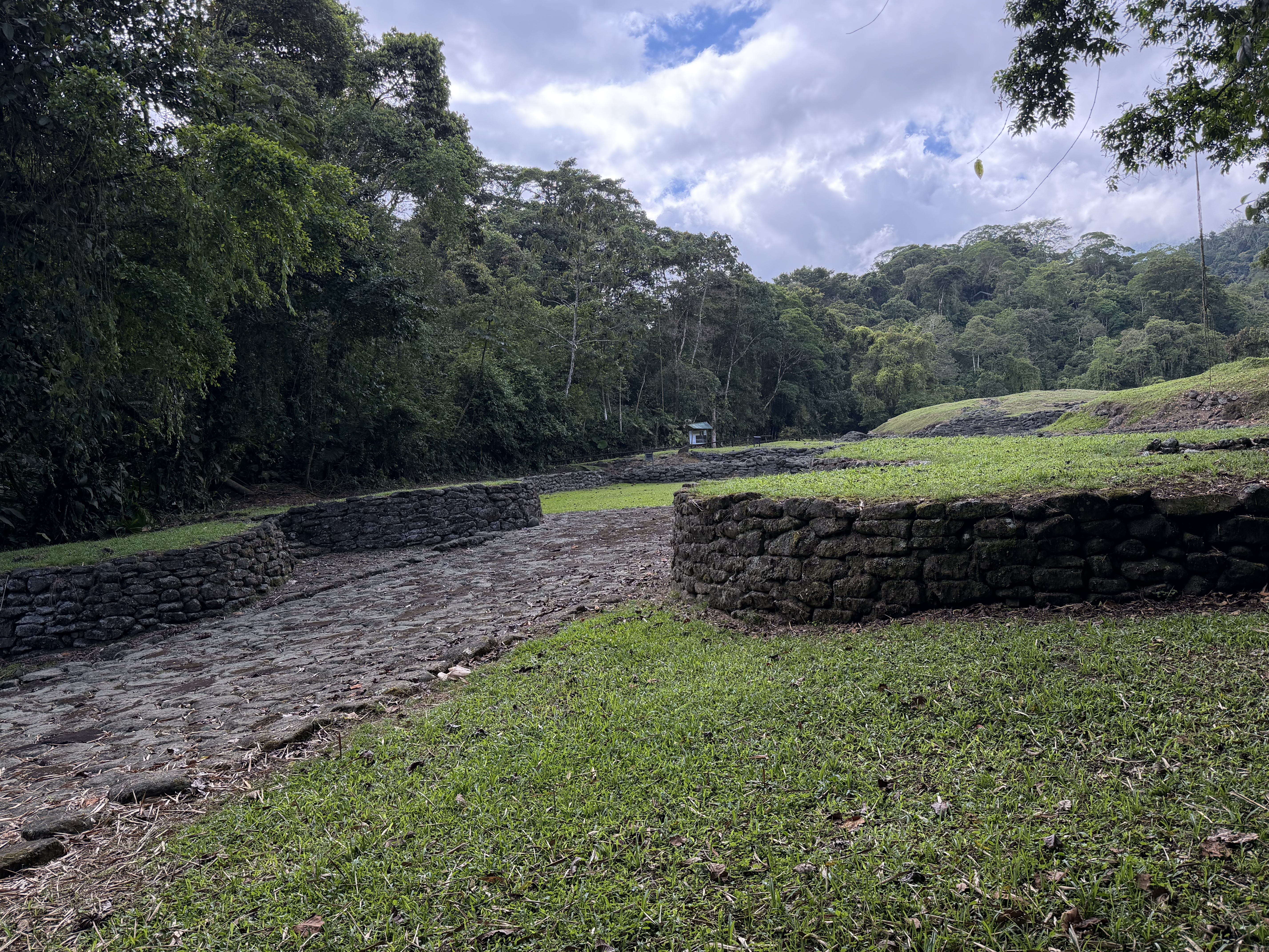 A view of an ancient stone structure surrounded by lush greenery and trees. The ground is covered in grass and stone pathways, leading to a prominent circular stone formation. In the background, there are additional stone remnants and a forested hillside under a partly cloudy sky. monumento nacional guayabo, parcelas