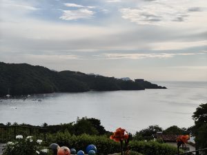 Coastal view with calm water, a few boats, green hills, and a partly cloudy sky.
