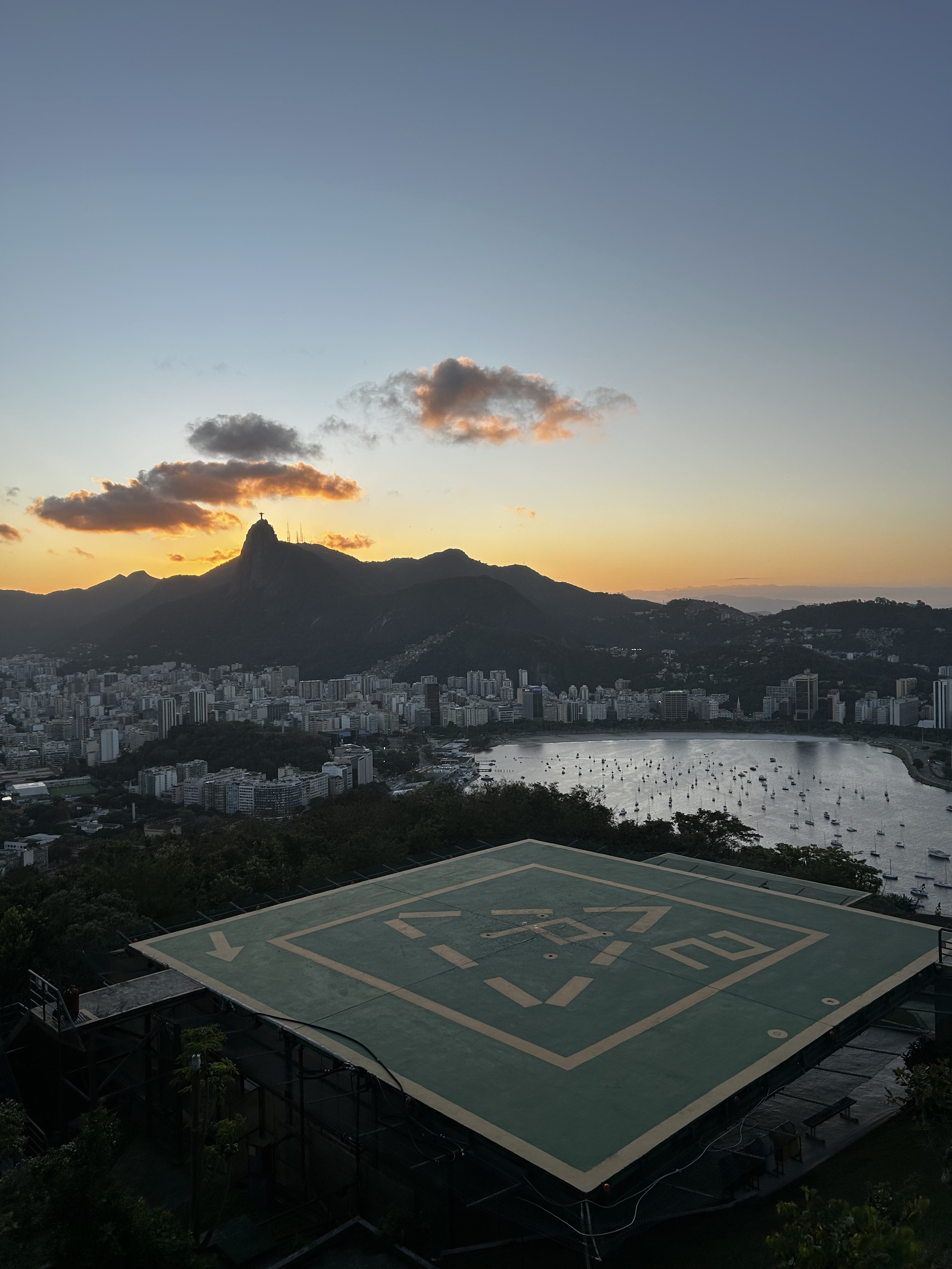 A panoramic view from a mountain overlooking a city at sunset. In the foreground, a helipad is visible, marked with yellow lines on a green surface. In the background, towering mountains blend into the skyline, with colorful clouds illuminated by the setting sun. The city below is dotted with buildings, some tall and modern, and features a body of water with boats anchored. The overall scene captures a serene and picturesque moment in nature and urban environment.