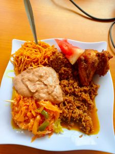A white square plate with spiced rice topped with fried chicken and watermelon, served with yellow pasta, cabbage salad, and brown sauce on a wooden table.