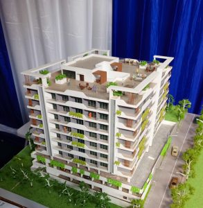 A white and grey multi-story building model is displayed on a green base representing grass. The model features multiple balconies with green miniature foliage, a rooftop terrace with small figures and furniture, and a ground-level area with a grey paved road and miniature cars. The background consists of blue and white fabric drapes.