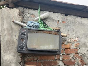 A rusty toaster oven placed on a weathered brick wall under a roof, stacked with rope, pipes, and plastic cups, showing a neglected scene.
