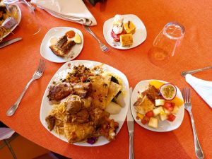 An orange tablecloth with plates of food, including rice, stew, pasta, chapati, fried fish, cakes, and sliced fruit.
