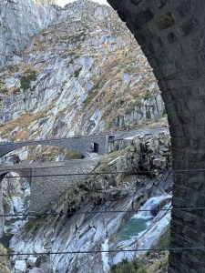 Gotthard bridges. A scenic view through a stone arch, showing a mountainous landscape with steep rocky cliffs and a winding road. The rocks are layered and colored in hues of gray and brown, with patches of green vegetation. Below, a river flows over rocks, reflecting shades of turquoise. 