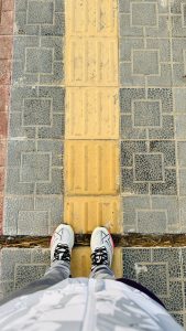 A top-down view of a person in white sneakers standing on a sidewalk with yellow tactile tiles, surrounded by gray patterned pavement.